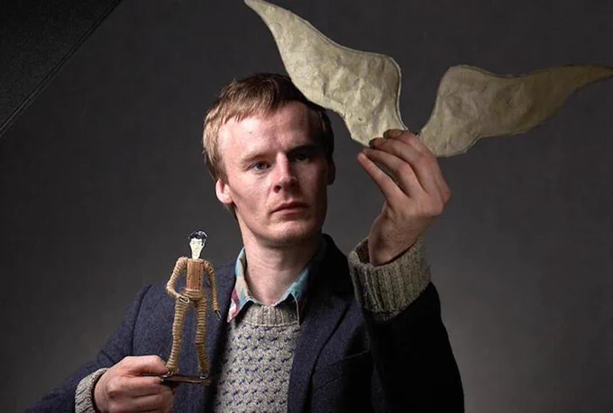 Photo of a performer wearing a knitted jumper and a jacket, holding a small figure of a man in one hand and a pair of paper wings in the other.