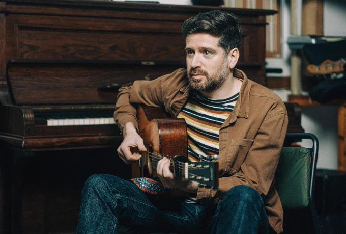 Photo of Kris Drever sitting next to a piano, playing an acoustic guitar.