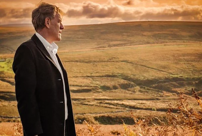 Photo of Martyn Joseph standing in a landscape of rolling hills and fields in golden sunlight.