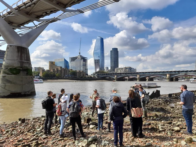 A group of people stood on the Thames foreshore. In the background is the Thames, with the underside of the Millennium Bridge and London skyline. #Mudlarking 