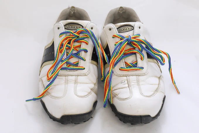 Image description - a pair of white and black FootJoy golf shoes, that look worn.  They have long rainbow coloured shoelaces tied in each shoe.