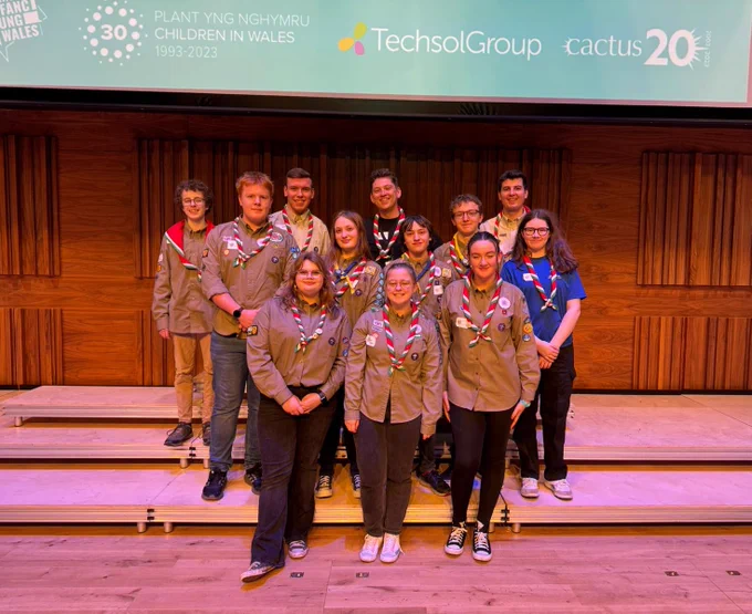 CEO of Children in Wales, Hugh Russell, is standing on a wooden stage with a group of young volunteers from the Welsh Scouts. 