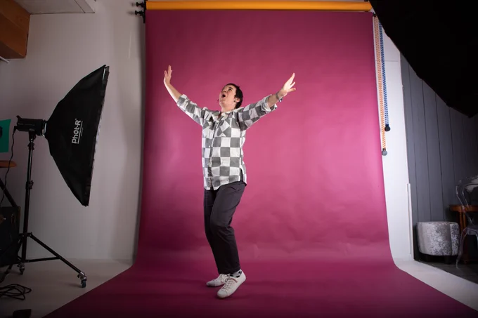 Photo of Katherina Radeva in a photography studio. She is wearing dark trousers, trainers and a black and white checked shirt, dancing on a dusky pink backdrop. 