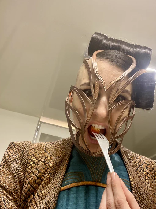 Jessica Boone with half head shaved wearing bronze snakeskin jacket and gilded mask looks down to camera biting strawberry