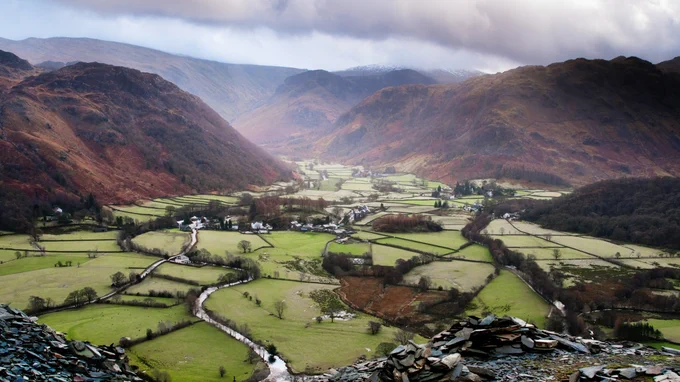 A valley with low cloud above. Green, farmed fields cross the land below, with trees and rugged land giving darker colours on the hills to the sides.