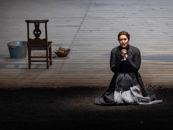 A scene from Janáček’s JENŮFA: A woman (Jenufa) kneels in the dirt, hands clasped in fervent prayer. Her brown hair is in a braided crown and she is wearing a simple black dress, floor-length with long sleeves and a high neckline, with a light blue printed cotton apron. To her left is a wooden chair, a metal tub, and a bowl of potatoes. Photo credit: Michael Brosilow