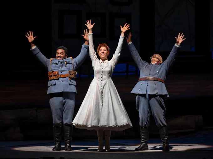 A scene from Donizetti's The Daughter of the Regiment: Three people stand with arms raised over their heads with big smiles. The woman in the middle (Marie) has short, red hair and is wearing a light blue pinstriped dress with buttons down the front and a while collar. The men on either side (Tonio and Sulpice) wear blue wool military uniforms with brown leather belts and black knee-high boots.  Photo credit: Michael Brosilow