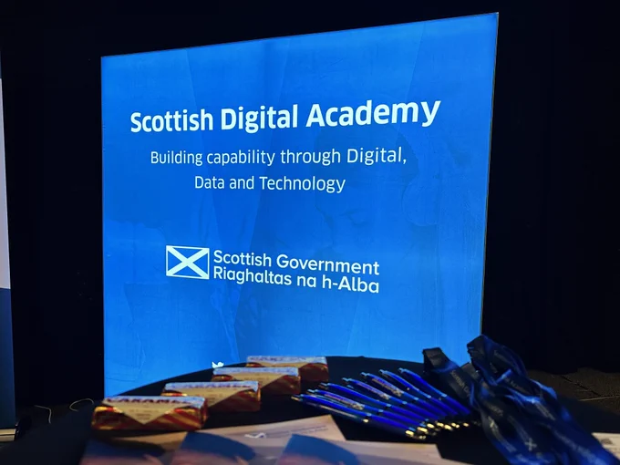 A blue light stand with white words Scottish Digital Academy. There is a table with pens and leaflets.