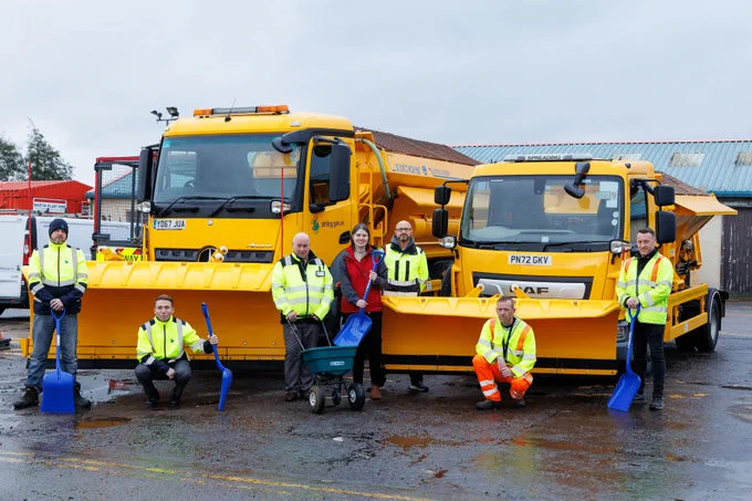 A team of council road workers, in yellow high vis vests and with shovels and salt barrows, stand either side of a women in red waterproof clothing and in front of two Gritter machines.