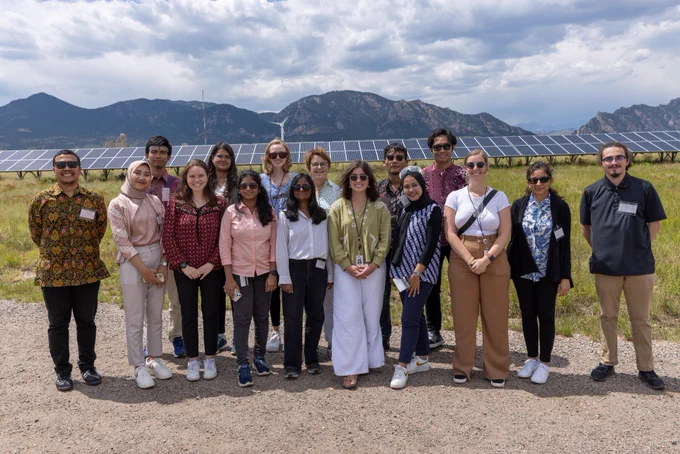 A group of people stand in front of the solar panels on NREL's flatirons campus. The mountains are in the background.