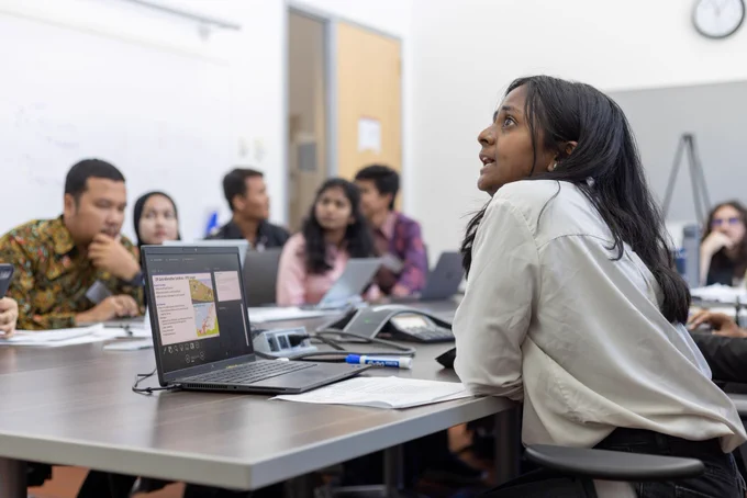 A group of people sit at a computer. The focus is on one woman who has her laptop open and is talking to someone off screen.