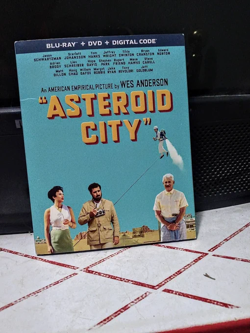 Blu-ray of asteroid city shows three white people standing in front of a desert town with a kid flying with a jet pack in the light blue sky.