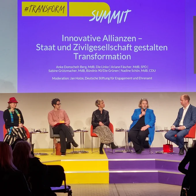 Three people are having a conversation on stage at the ‘transform_d Summit’. The title of the discussion can be read in the background: "Green but unfair? - What is needed for a fair climate transition?".