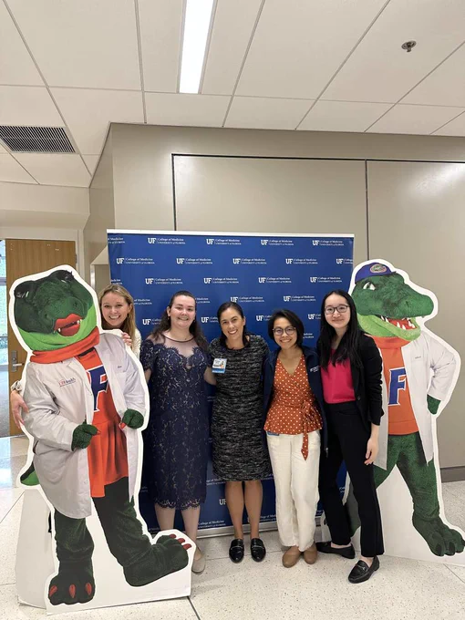 Five female students in front of a blue backdrop with the U-F College of Medicine logo and smile while standing next to cardboard cutouts of Alberta and Albert Gator mascots wearing white coats.