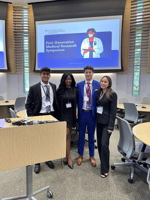 Four students stand in a lecure hall and smile at the camera in front of a projector screen that reads, "First-generation medical research symposium."