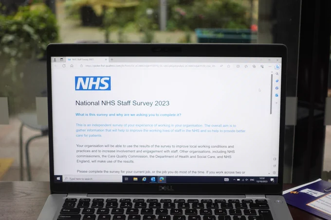 An opened laptop on a table shows the front page of the Staff Survey 2023 website. The background shows a small hospital garden. 