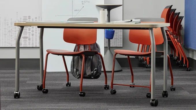 The Steelcase Learning Agree table and red Steelcase Learning Tenor chairs on wheels are featured in an active learning space. Additional Tenor chairs are stacked in the back of the room along with a freestanding Whiteboard for collaboration.