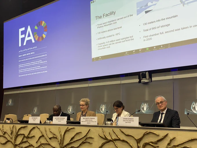 Row of two men and two women sitting at a podium. One woman presenting in front of a screen.