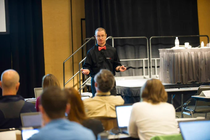 ServiceNow employee Chuck Tomasi presents to a small group of people sitting at their laptop computers at a Knowledge conference. He is wearing a red bowtie and black shirt.