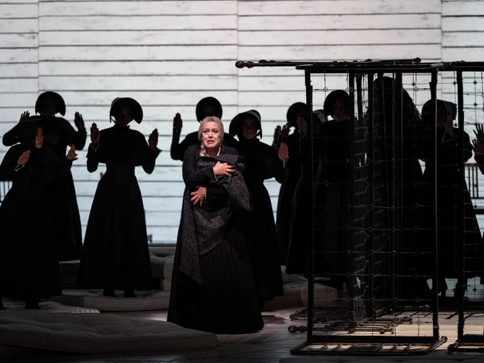 Soprano Nina Stemme as Kostelnička clutches a baby in a blanket. She is wearing a floor-length black dress with long sleeves and a high collar. Next to her, you can see metal bed frames. In the background, women in black dresses and bonnets look on menacingly.