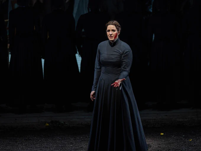 Soprano Lise Davidsen as Jenůfa, stands in a black floor-length dress with long sleeves and a high collar. One hand is outstretched, covered in blood, as is her cheek which was slashed with a knife.