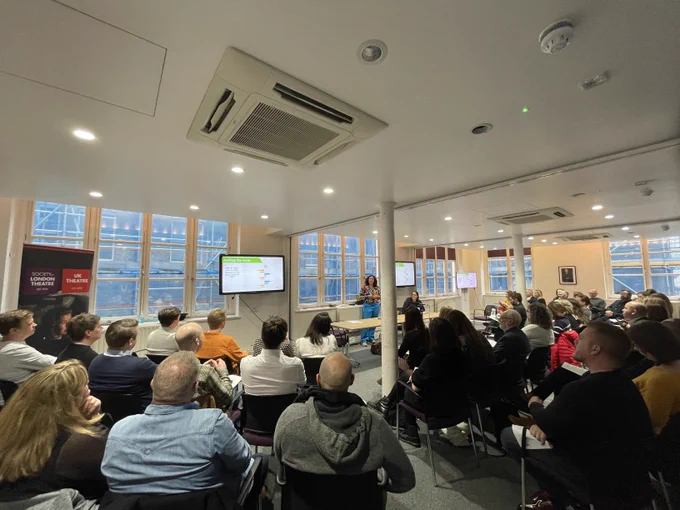 A large, bright meeting room viewed from the back, filled with people all paying attention to the person speaking at the front of the room.
