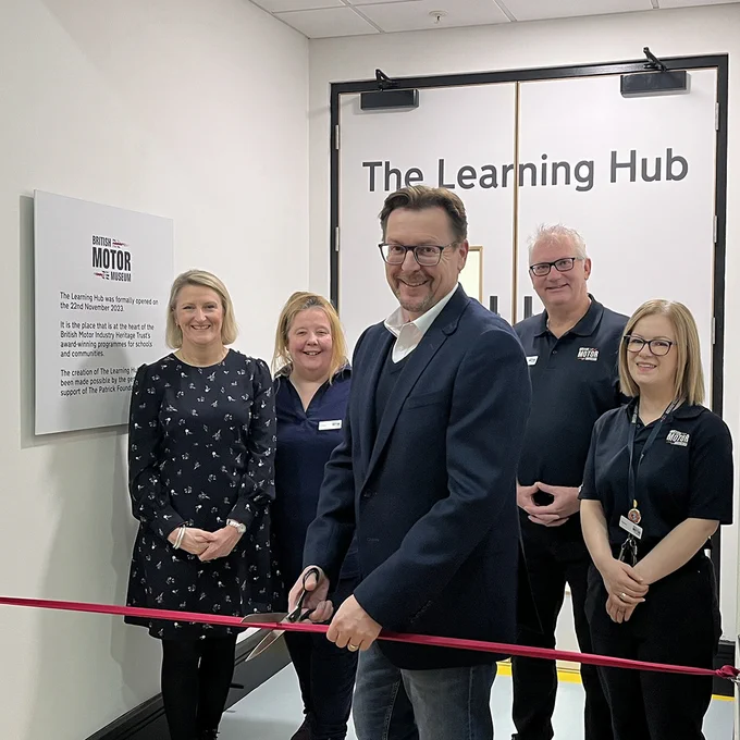 The Patrick Foundation and the British Motor Museum Learning & Enagement Team standing in front of the doors to The Learning Hub. In the centre of the photo, Julian is holding and preparing to cut a red ribbon.
