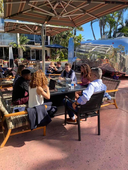 A picture of Julius Mbura wearing a black shirt and maroon pants. He is seated outside with other people around a square table, and they’re having a conversation together.