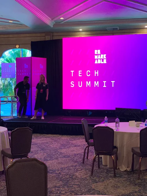 A picture of a stage that has two people dressed in black next to a large screen. The large one has a pink and purple background, and it’s written Remarkable Tech Summit.