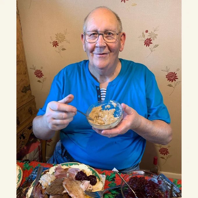 A photo of Gerald Stratford wearing a blue shirt, sitting at a dinner table holding a bowl and a spoon.