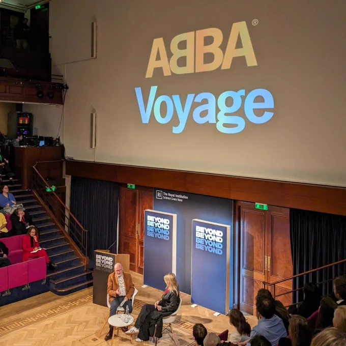 A man and woman sit on the stage at the Royal Institution with the BEYOND branding behind them and 'ABBA Voyage' in nig lettering on the screen above them.
