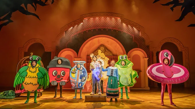 7 children in costumes of objects (hoover, vase, inflatable flamingo etc) stand in a line and sing. Behind them is a pink marble concierge desk at a htoel.
