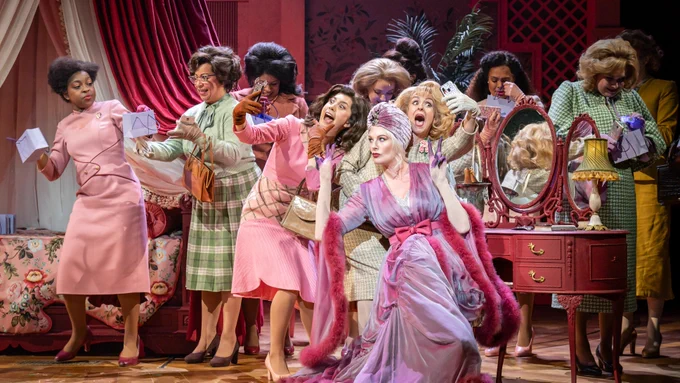 A woman in a purple dressing gown and head scar with pink feather lining sits on a stool at a dressing table. A large group of women in 1950s esque pastel clothes stand around her. Two take a selfie with her. There is a grand four poster bed behind them. The women hold small purple gift bags.