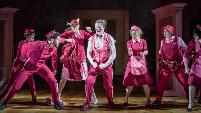 Seven people in pink hotel uniform for maids and bell boys etc stand in a line. The man in the centre has ripped sleeves and war paint on his face. Pink door frames stand behind them.