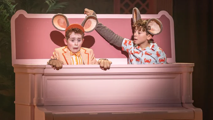 Two young boys with large fake mouse ears and grey fur arms with shirts over the top pop out from the top of a pink piano. One looks terrified the other calms him.