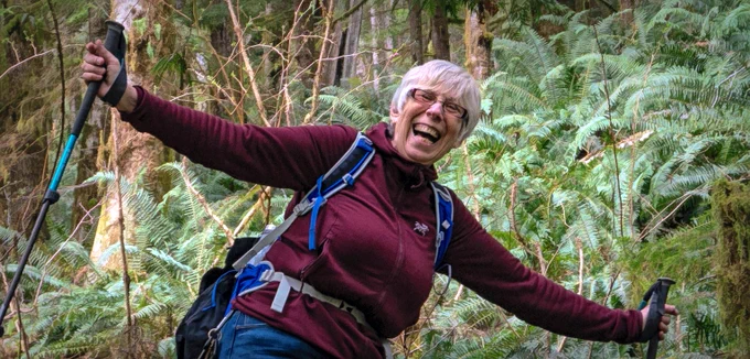 A woman with short gray hair carrying hiking poles and a daypack gives a huge smile as she hikes past green ferns
