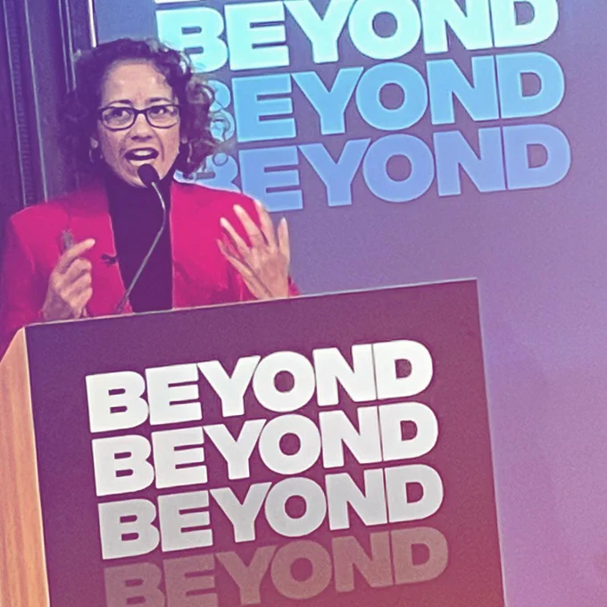 Broadcaster Samira Ahmed, a woman with dark curly hair and glasses, speaks animatedly behind a lectern, with the BEYOND logo behind her and on the lectern.