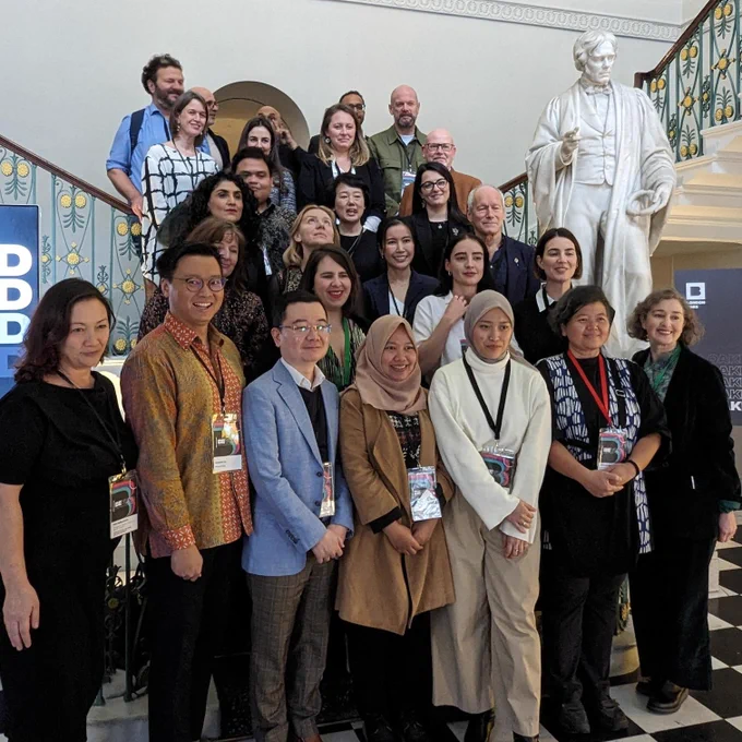 A group of 27 people of different nationalities that make up the British Council delegation stand on the staircase at the Royal Institution, smiling and posing for the photo. A marble statue is next to them.