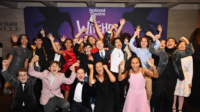 A large group of children in suits and pretty dresses all stand and cheer. Behind them is a backdrop that is purple with two black illustrated hands and white lettering reads 'National Theatre' 'The Witches'.