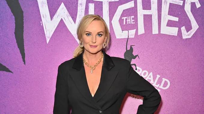 A woman with blonde hair in a black suit jacket and gold jewellery smiles at the camera.  Behind her is a backdrop that is purple with two black illustrated hands and white lettering reads 'National Theatre' 'The Witches'.
