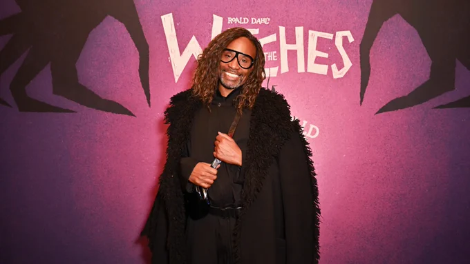 A person in a black shirt, trousers and fur coat with long brown hair smiles at the camera.  Behind them is a backdrop that is purple with two black illustrated hands and white lettering reads 'National Theatre' 'The Witches'.