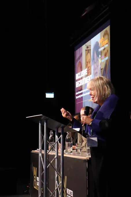 This is an iamge of Dame Julia Cleverdon's side profile. She is standing at a podium, wearing a black coat with a blue collar holding a mic in her hand. 
