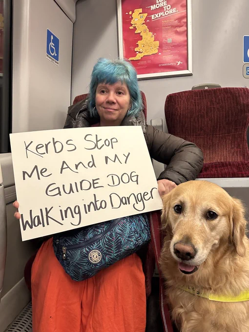 Picture of Sarah Leadbetter and Nellie her Guide Dog sitting on a train holding a board with the words ‘Kerbs stop me and my Guide Dog walking into danger’.