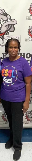 Leyla is wearing a purple t-shirt that says ESOL Squad in big, colorful letters. She is wearing black pants and black shoes, and is standing in front of a wall at her school that has their school mascot, a bulldog, on it.