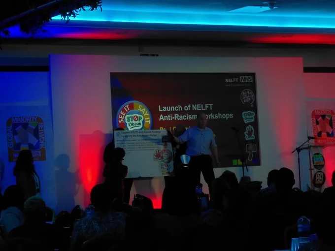 NELFT CEO Paul Calaminus on stage at the Global Majority Network conference introducing NELFT's Anti-Racism pledge. He's standing in front of an oversized pledge card as senior leaders line up to add their signatures.