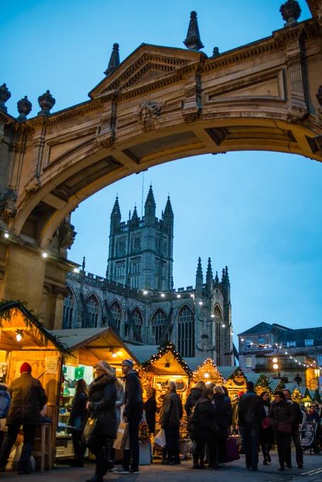 People are walking around the Bath Christmas Market chalets. In the background you can see Bath Abbey and the stone arch above.