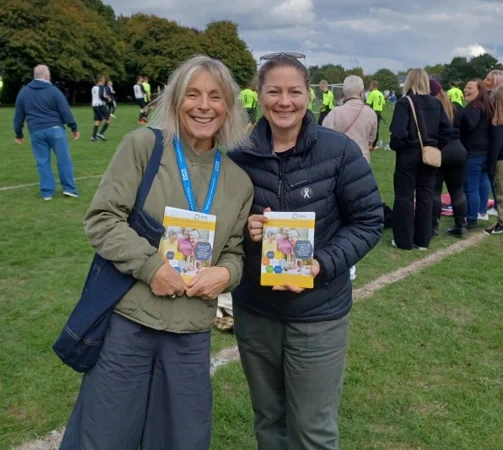 Two ladies from the Community Care Transformation programme are standing in a sports field smiling and holding some leaflets.