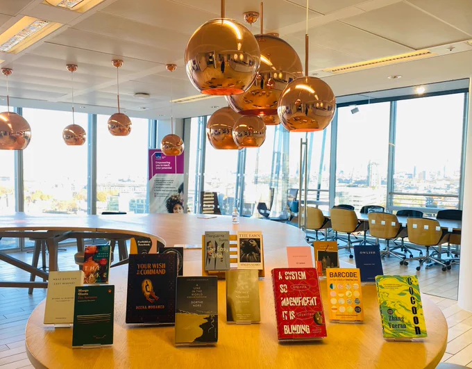 The eight shortlisted titles for the Warwick Prize for Women in Translation are stood in a row on a wooden table. Behind are the eight long listed titles for the prize also in a row. There are light fittings above the table and in the background is a glass wall with a view over London. 