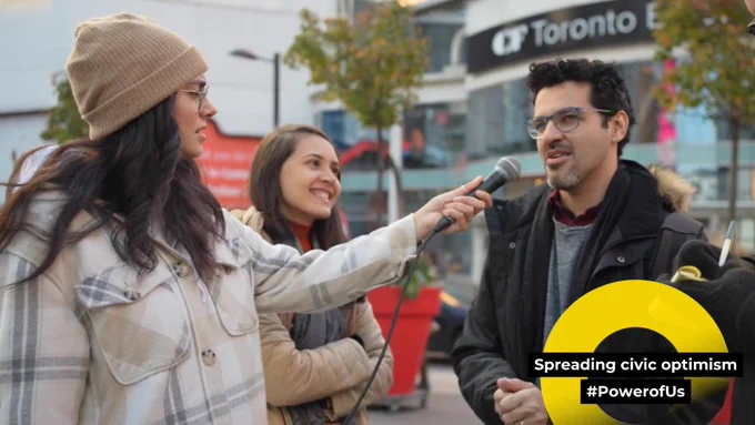 Three people standing outside in a public square. Woman wearing a toque and jacket is holding a microphone out to a man who is talking. Woman in background is smiling and looking at the man.  