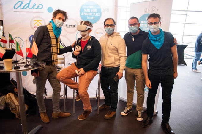 A group of people posing for a photo. One person is sitting in a tall chair with a VR Headset on. 